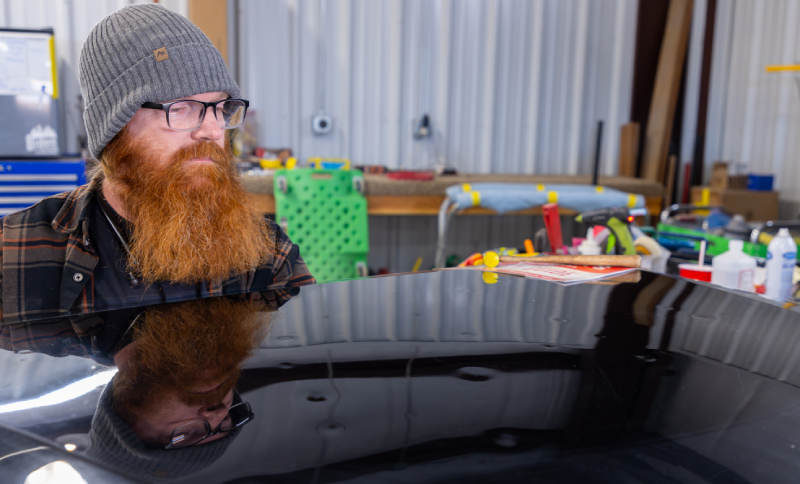 A person with a beard and beanie looks at the polished surface of a car hood in a workshop.