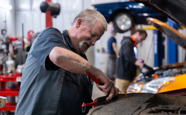 A mechanic with a mustache works on a car engine with a red tool. In the background, another mechanic is visible near a vehicle on a lift.