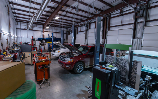 Auto repair shop interior with several vehicles, tools, and equipment. A red SUV has its hood open, with other cars on lifts and various machinery around the room.