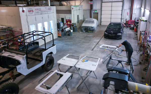 Auto body shop interior with various vehicle parts being repaired and painted. Worker sanding a car door. Paint booth and two vehicles are visible.