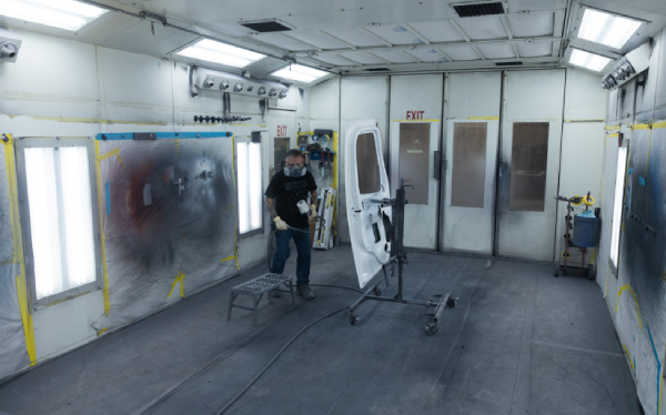 A person in protective gear is working with a spray gun in an industrial paint booth, painting a car door. Safety equipment and walls are visible in the background.