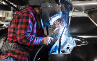 A person in a welding helmet is welding the frame of a vehicle in a workshop. Sparks are visible.