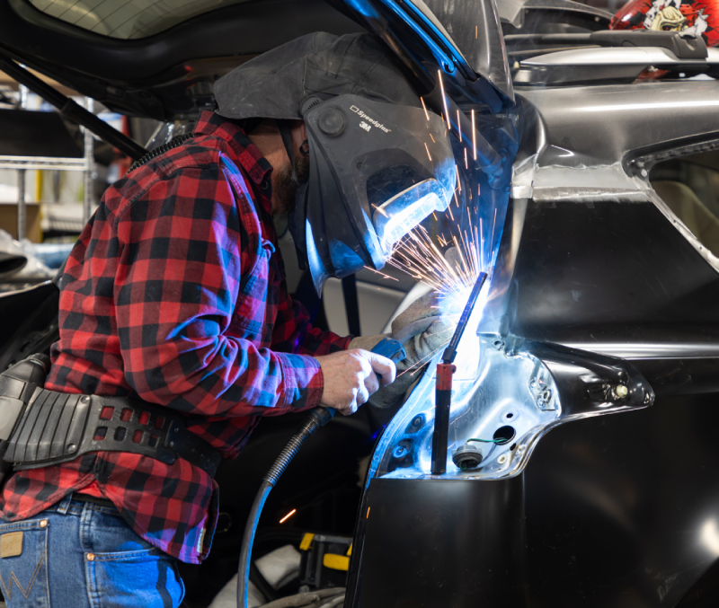 A person in a welding helmet is welding the frame of a vehicle in a workshop. Sparks are visible.