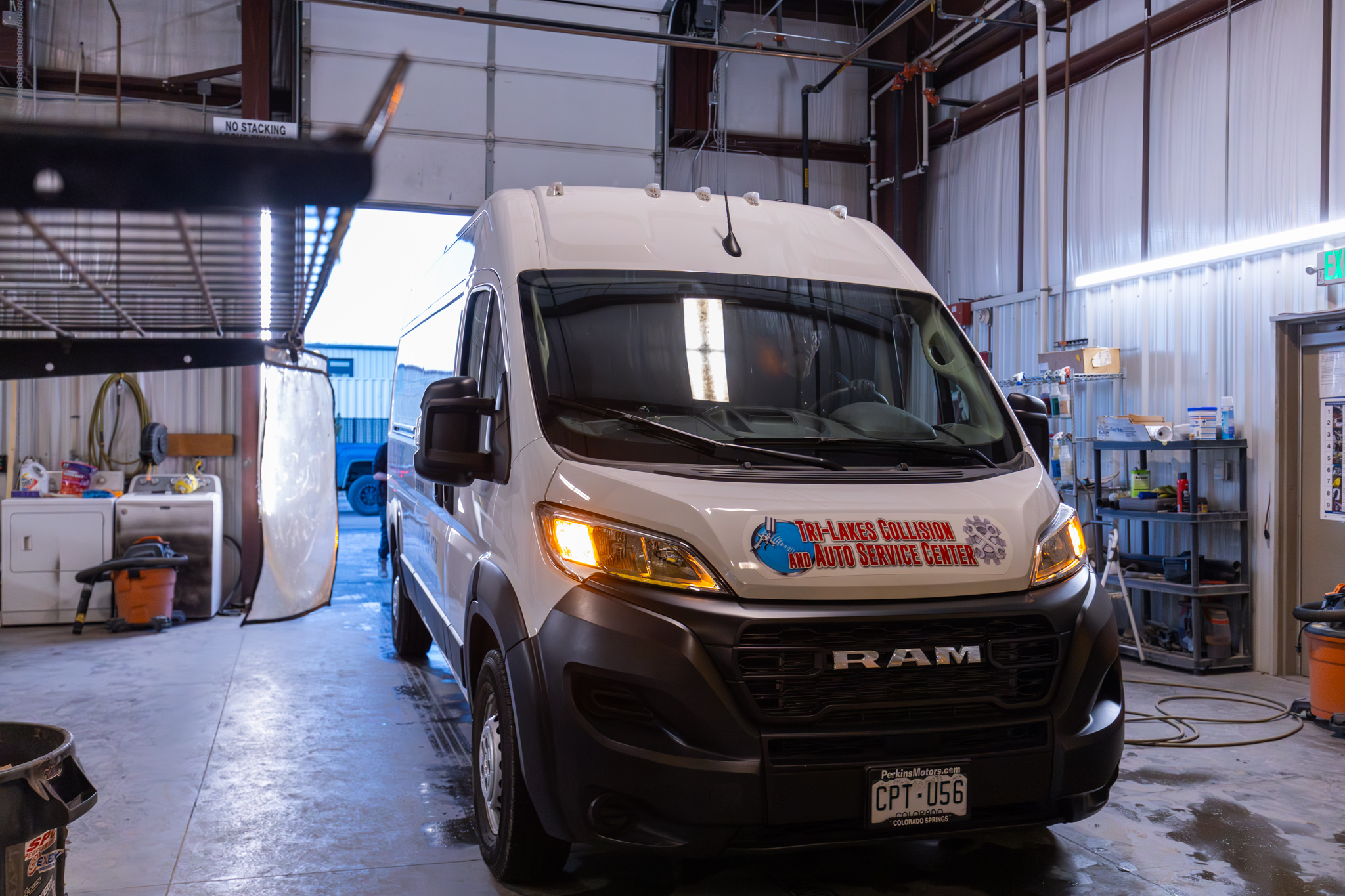 A white RAM van with "Tri-Lakes Collision and Auto Service Center" signage is parked inside a garage, surrounded by auto repair tools and equipment for services like auto glass replacement.