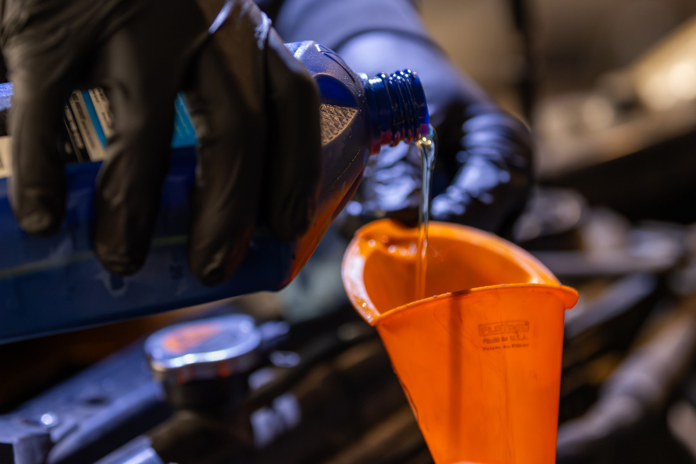 A person wearing gloves carefully pours blue fluid into an orange funnel on a car engine, showcasing the meticulous art of auto restoration.