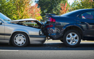 A silver car has rear-ended a dark blue car on a road, causing visible damage to the front of the silver car and the back of the blue car.