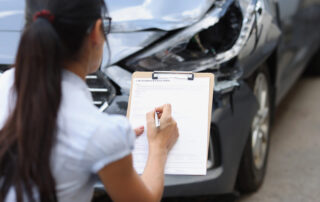 A woman fills out a car insurance claim form on a clipboard while inspecting a damaged vehicle with a dented front and broken headlight.