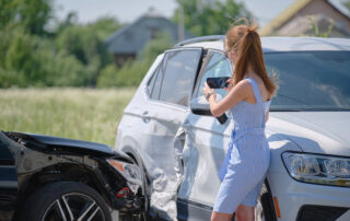A woman takes photos of car damage with her phone after a collision between a white SUV and a black vehicle on a sunny day.